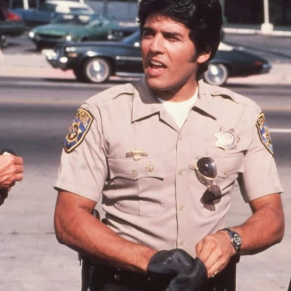 A man in a beige sheriff's deputy uniform with badges, sunglasses, and a black glove, standing outdoors with cars blurred in the background.